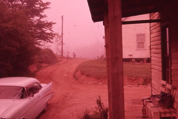 vintage view of people walking down a dirt road. There's a vintage car and an old building